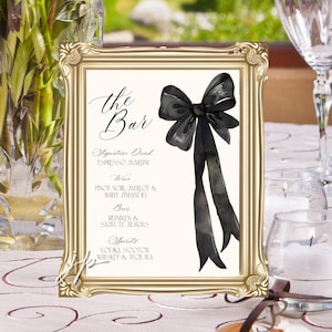 May include: A framed sign with the text "the Bar" and a black bow illustration. The sign lists drink options: Espresso Martini, Pinot Noir, White Zinfandel, Heineken, Samuel Adams, Vodka, Scotch, Whiskey, and Tequila. The frame is gold.