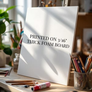 May include: A white rectangular foam board standing upright on a wooden surface. The board features black text that reads PRINTED ON 3/16" THICK FOAM BOARD. The surface includes various art supplies like colored pencils and a glue stick.