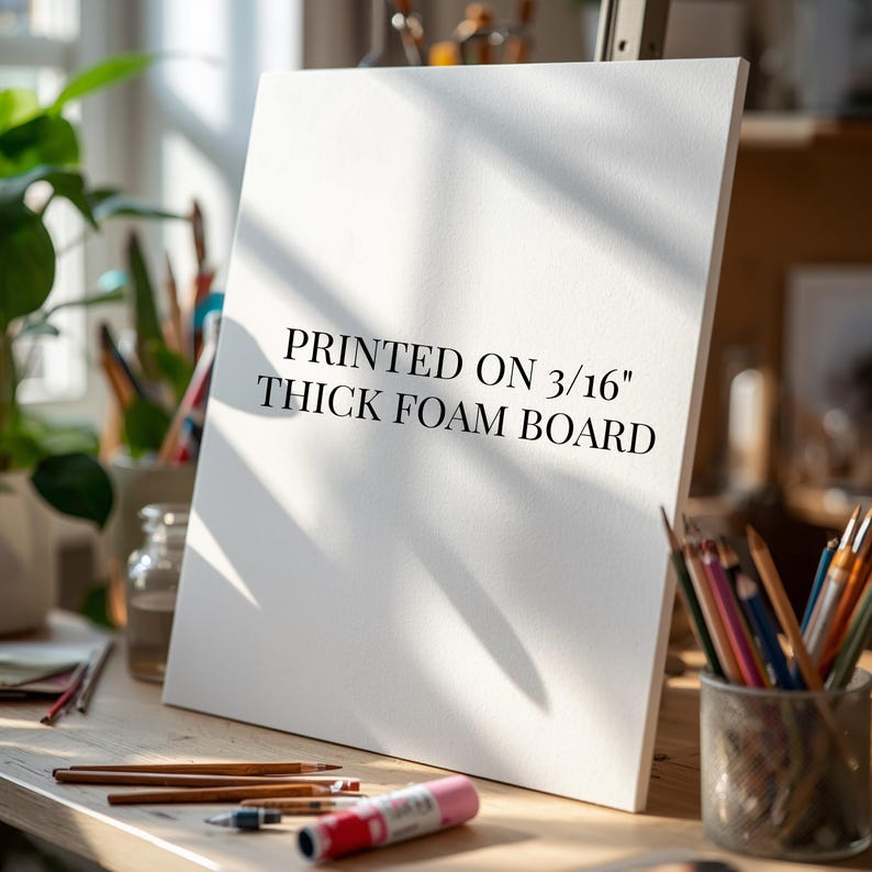 May include: A white rectangular foam board standing upright on a wooden surface. The board features black text that reads PRINTED ON 3/16" THICK FOAM BOARD. The surface includes various art supplies like colored pencils and a glue stick.