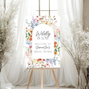 May include: A rectangular sign featuring a white arched center with the text Wildly IN LOVE WELCOME TO Samantha's BRIDAL SHOWER, surrounded by a colorful watercolor floral border of blue, orange, pink, and yellow wildflowers and green foliage.