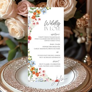 May include: A white wedding menu card with floral watercolor accents. The card features the words "Wildly in Love" and a menu with appetizers, main course, and dessert options. The card is set on a decorative plate.
