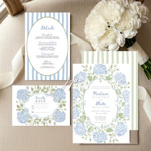 May include: A wedding stationery set featuring a rectangular invitation, a details card, and an RSVP card. The invitation and RSVP card display blue hydrangea illustrations with green leaves. The details card has blue vertical stripes.