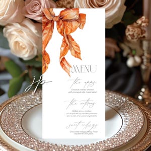 May include: A wedding menu card with an orange watercolor bow design. The menu includes appetizers, entrees, and desserts. The card is placed on a decorative plate with a floral background.