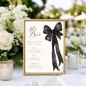 May include: A gold-framed sign featuring a black watercolor bow illustration and the text: the Bar, Signature Drink Espresso Martini, Wine Pinot Noir, Merlot & White Zinfandel, Beer Heineken & Samuel Adams, Spirits Vodka, Scotch, Whiskey & Tequila.