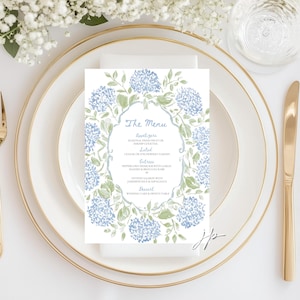 May include: A rectangular menu card featuring a watercolor illustration of blue hydrangea flowers and green leaves forming a decorative border. The text reads The Menu, Appetizers, Salad, Entrees, and Dessert with listed food items in blue ink.