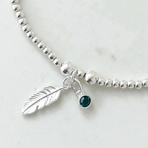May include: A silver beaded bracelet with a silver feather charm and a dark green gemstone charm.