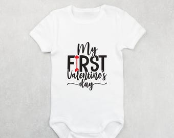 May include: White baby onesie with black text that reads "My First Valentine's Day" with a red arrow in the word "First".