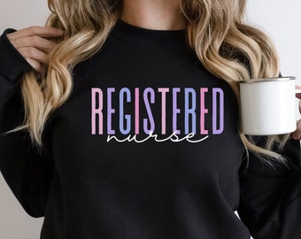 Puede incluir: Sudadera negra con las palabras "REGISTERED nurse" en un degradado de rosa, morado y azul. Una taza blanca se sostiene en la mano. La sudadera es de estilo cuello redondo.