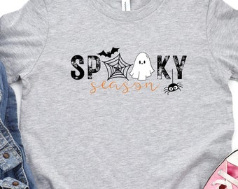 Könnte beinhalten: Ein graues, kurzärmeliges T-Shirt mit dem schwarzen Schriftzug SPOOKY, einem Spinnennetz, einem Geist und einer Fledermaus sowie dem orangefarbenen Wort season und einer kleinen schwarzen Spinne. Eine blaue Jeansjacke und rosa Sneaker.