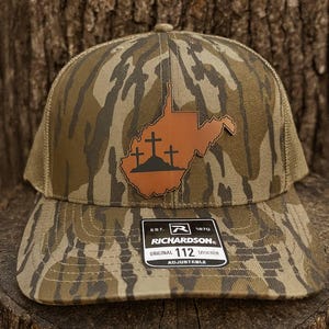 May include: Camouflage pattern trucker hat with a brown leather patch featuring the outline of West Virginia and three crosses. The hat has a curved brim and a mesh back. The Richardson brand label is visible.