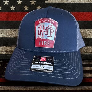 May include: A navy blue trucker hat with a Hamlin Fire patch. The patch is red and silver with the word "FIRE" at the bottom. The hat has a mesh back and an adjustable strap. The background is an American flag.