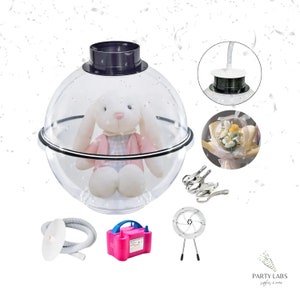 May include: A clear plastic sphere with a black rim, containing a stuffed white bunny. The sphere is part of a set for creating balloon bouquets, which also includes a pink electric air pump, a clear plastic tube, a silver metal stand with a fan, and silver clips.