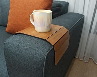 May include: A wooden sofa arm tray with a coffee mug on top. The tray is rectangular and fits over the arm of a gray sofa. An orange pillow rests against the sofa back.