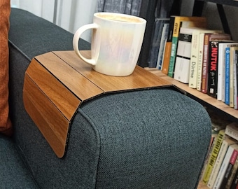 May include: A wooden sofa arm tray with a cup of coffee on it. The tray is made of wood and has a curved design to fit over the arm of a sofa. An orange pillow is on the sofa. A bookshelf with books is in the background.