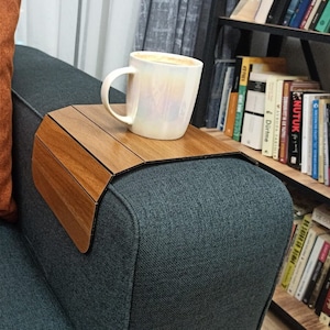 Könnte beinhalten: Ein hölzernes Sofatablett mit einer Tasse Kaffee darauf. Das Tablett ist aus Holz gefertigt und hat ein gebogenes Design, um über die Armlehne eines Sofas zu passen. Ein orangefarbenes Kissen befindet sich auf dem Sofa. Ein Bücherregal mit Büchern befindet sich im Hintergrund.