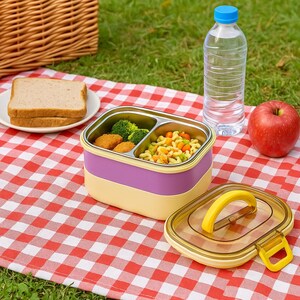 Op de afbeelding: Een lunchbox met twee niveaus met een roestvrijstalen binnenkant, met broccoli, nuggets en pasta. De doos is paars en geel, met een doorzichtig deksel en een gele handgreep. Een fles water, een rode appel en een bord brood staan in de buurt.