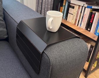 Può includere: Un vassoio per bracciolo di divano in metallo nero con una tazza bianca sopra. Il vassoio è rettangolare con un pannello laterale con linee orizzontali. Il bracciolo del divano è grigio e una libreria è visibile sullo sfondo.