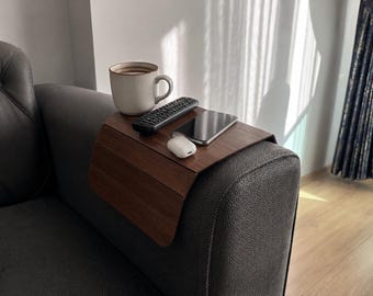 Può includere: Un vassoio in legno marrone scuro per bracciolo del divano con design flessibile a doghe, posizionato sopra il bracciolo di un divano grigio testurizzato. Il vassoio sostiene una tazza in ceramica bianca, un telecomando nero, uno smartphone e un astuccio.
