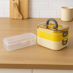 May include: A two-tiered yellow and cream-colored lunch box with a gray handle and latches. A clear rectangular container sits to the left. The lunch box is on a wooden surface.