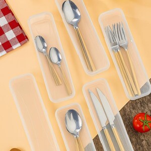 May include: A collection of stainless steel silverware with gold-colored handles, including spoons, forks, and knives, presented in clear plastic cases. A tomato, salt shaker, and olive oil bottle are also visible.