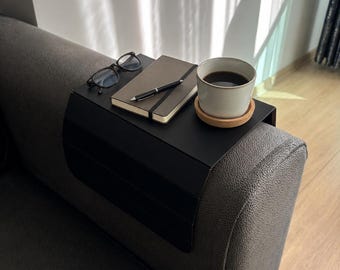 Può includere: Un vassoio da bracciolo per divano in nero opaco con una superficie piana che poggia sul bracciolo. Sul vassoio sono appoggiati un paio di occhiali con montatura nera, un taccuino grigio chiuso con elastico nero, una penna nera e una tazza bianca.