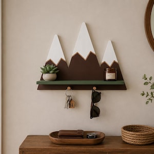 Può includere: Una mensola da parete a forma di montagna con tre cime, punte bianche e basi marroni. La mensola presenta un ripiano orizzontale verde con due ganci in legno che sostengono un mazzo di chiavi e un paio di occhiali da sole. Una pianta e una candela.