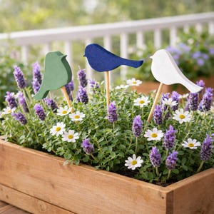 May include: Three wooden bird garden stakes in green, blue, and white, set in a wooden planter box filled with lavender and daisies. The birds are on wooden dowels, enhancing the garden's decorative appeal.