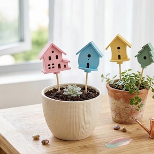 Puede incluir: Cuatro estacas decorativas para plantas con formas de madera en miniatura: una casa rosa, una casa azul, una casa amarilla y un árbol verde. Cada forma está montada sobre un palo de madera fino, diseñado para insertarse en macetas.