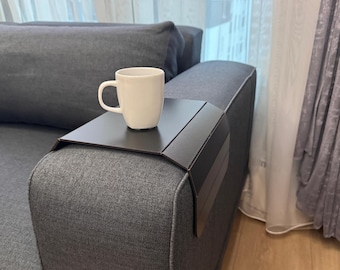 Può includere: Un vassoio per bracciolo del divano in metallo marrone scuro con una tazza bianca sopra. Il vassoio è progettato per adattarsi al bracciolo di un divano grigio. La tazza ha un manico curvo e un design semplice e pulito.
