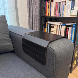 May include: A black metal sofa arm tray with a rectangular top and a side panel with horizontal lines. The tray is placed on the arm of a gray sofa. The background includes a bookshelf filled with books.
