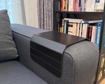 Può includere: Un vassoio per bracciolo del divano in metallo nero con un piano rettangolare e un pannello laterale con linee orizzontali. Il vassoio è posto sul bracciolo di un divano grigio. Lo sfondo include una libreria piena di libri.