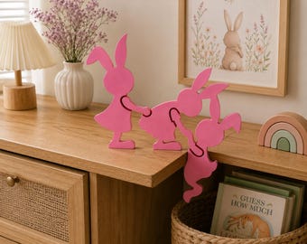 May include: A set of three interlocking pink wooden rabbit figures. The rabbits are connected by their hands, with two standing on a flat surface and the third hanging over the edge. Each rabbit has long ears and a simple silhouette design.