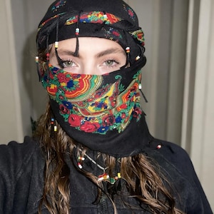May include: Black headscarf with a colorful floral pattern covering the lower face. The head covering is adorned with beaded tassels. The floral design includes red roses, blue and yellow flowers, and green leaves.