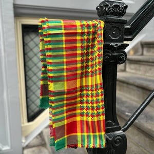 May include: A vibrant scarf with a plaid design in red, yellow, green, and brown is draped over a black metal railing. The scarf features a fringed edge and a houndstooth pattern on one side.