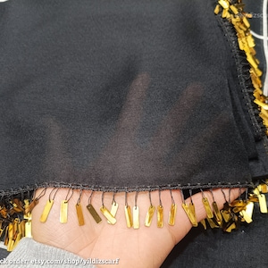 May include: Black scarf with gold rectangular fringe. The scarf is made of a sheer fabric and has a black border. The background is a black and white checkered pattern. The Etsy shop name is visible in the image.
