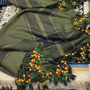 May include: Olive green plaid scarf with decorative fringe and beaded accents in red, yellow, and green. The scarf has a classic plaid pattern with black and tan stripes. The fringe is dark green and the beads are small and colorful.