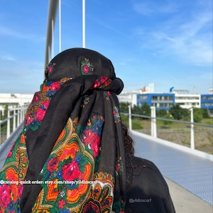 May include: A black headscarf with a colorful floral pattern. The scarf is tied around the head and the ends are tucked in. The scarf is worn by a person standing on a bridge. The text "@yildizscarf" is visible in the bottom right corner of the image.