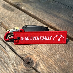 May include: Red rectangular keychain tag with the text "0-60 EVENTUALLY" and a speedometer graphic. The tag is attached to a red carabiner clip, keys, and a black car key fob. The items are on a wooden surface.