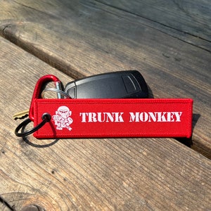 May include: A red fabric keychain tag with the words "TRUNK MONKEY" in white block letters. A cartoon monkey graphic is on the left. A black car key and a key are attached to the tag with a red carabiner clip.