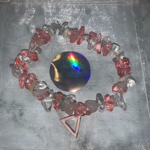 Könnte beinhalten: Ein rosa und klares Kristall-Armband mit einem rosa Dreiecks-Anhänger. Das Armband liegt auf einem rosa und weißen Netzbeutel mit einem Aufkleber, auf dem "Vielen Dank für Ihren Einkauf bei uns" steht.
