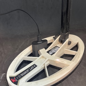 Könnte beinhalten: Ein weißer Metalldetektor mit einem schwarzen Griff und einem schwarzen Ladekabel, das in das Batteriefach eingesteckt ist. Der Detektor hat eine große, runde Suchspule mit dem aufgedruckten Text "HF COIL GOLD PROSPECTING".