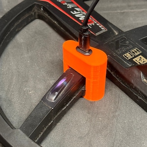 Könnte beinhalten: Ein orangefarbener 3D-gedruckter Adapter für einen Metalldetektor. Der Adapter ist mit einem schwarzen Kabel verbunden und am Arm des Metalldetektors befestigt.