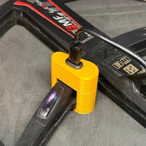 Könnte beinhalten: Ein gelber Kunststoff-Ladeständer für einen Metalldetektor. Der Ständer hat ein schwarzes Kabel daran befestigt. Der Ständer ist mit einem schwarzen Metalldetektor verbunden, auf dem der Text "METAL DETECTORS" und "28 CM" aufgedruckt ist.