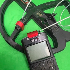 Puede incluir: Un detector de metales negro con una etiqueta roja y blanca que dice "XP DEUS II". El detector tiene una caja de control negra con una pantalla y botones. La caja de control está conectada a la bobina mediante un cable blanco.