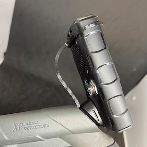 Könnte beinhalten: Ein schwarzer Metalldetektor mit einem grauen Griff und einem schwarzen Steuerkasten, der oben angebracht ist. Der Steuerkasten hat einen schwarzen Gurt, der daran befestigt ist. Der Griff trägt den Schriftzug "XP METAL DETECTORS".