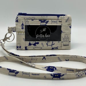 May include: A white fabric ID badge holder with a blue zipper, a clear ID window, and a blue and white patterned lanyard. The holder has a blue and white cat design with the text "stella bee handmade".