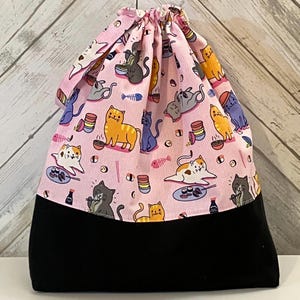 May include: A drawstring bag with a pink upper section featuring a repeating pattern of cartoon cats, sushi, and fish bones. The lower section is solid black. The bag is gathered at the top with a drawstring.