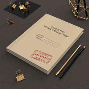 May include: A beige notebook titled "PLANS FOR WORLD DOMINATION" with a "TOP SECRET" stamp. Gold and black pencils and gold binder clips are arranged around the notebook on a dark gray surface.