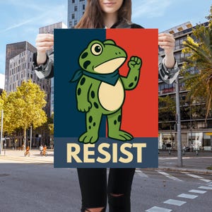 May include: A poster featuring a cartoon frog with a raised fist, wearing a blue bandana, against a red and blue background. The word "RESIST" is printed in a bold, sans-serif font at the bottom. The poster is held up in front of a city street.