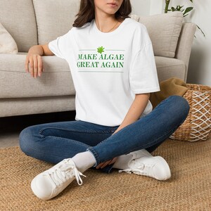 May include: A white short-sleeved t-shirt featuring the green text "MAKE ALGAE GREAT AGAIN" centered on the chest, accompanied by a small green graphic of an algae cluster above the text, paired with blue denim jeans and white sneakers.